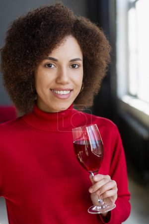 Foto de Retrato de una hermosa mujer afroamericana sonriente sosteniendo una copa de vino tinto mirando el cumpleaños de la celebración de la cámara en la fiesta en casa. Mujer feliz vistiendo rojo elegante cuello alto bebiendo alcohol - Imagen libre de derechos
