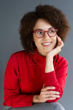Foto de Retrato de una sonriente mujer afroamericana confiada con gafas rojas y elegantes sentada en la oficina. Estudiante universitario exitoso inteligente en jersey de cuello alto rojo mirando hacia otro lado, concepto de educación - Imagen libre de derechos