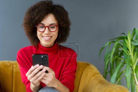 Foto de Sonriente mujer afroamericana sosteniendo el teléfono móvil viendo videos sentados en casa. Mujer moderna con gafas rojas usando aplicación móvil, compras en línea, pedidos de alimentos. Tecnología, banca móvil - Imagen libre de derechos
