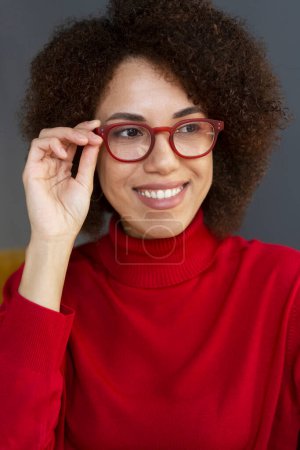 Foto de Retrato sonriente mujer afroamericana con elegantes anteojos rojos y suéter de cuello alto rojo mirando hacia otro lado aislado en el fondo. Concepto de visión - Imagen libre de derechos