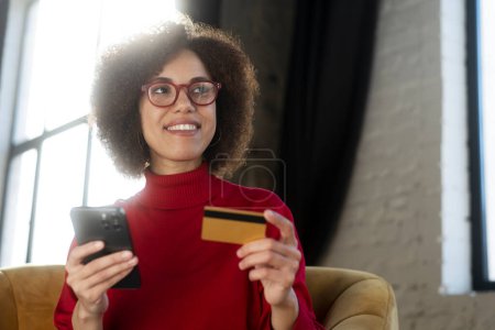 Téléchargez les photos : Femme afro-américaine souriante tenant des achats par carte de crédit en ligne, commandant de la nourriture sur le site Web assis à la maison. Le succès freelance portant des lunettes rouges élégantes reçoivent le paiement, travaillant en ligne - en image libre de droit