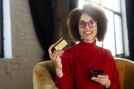 Foto de Sonriente mujer afroamericana sosteniendo tarjeta de crédito y teléfono inteligente, utilizando la aplicación móvil, compras en línea, pidiendo comida de casa. El freelancer exitoso y con estilo recibe el pago. Compra, concepto de venta - Imagen libre de derechos