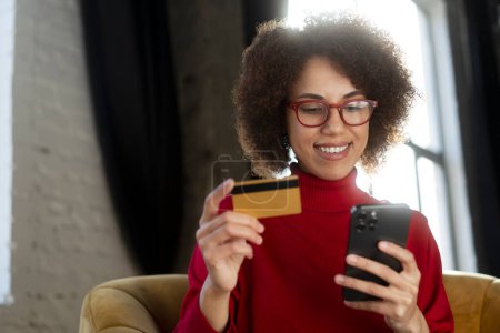 Photo for Smiling successful African American woman freelancer holding credit card and smartphone receive payment, working from home. Modern stylish female using mobile app shopping online, ordering food - Royalty Free Image