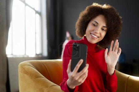 Téléchargez les photos : Portrait souriant femme afro-américaine tenant la communication par téléphone mobile en ligne, saluant la main, ayant un appel vidéo assis sur un fauteuil à la maison. Influenceur enregistrement vidéo à l'aide de smartphone. Médias sociaux - en image libre de droit