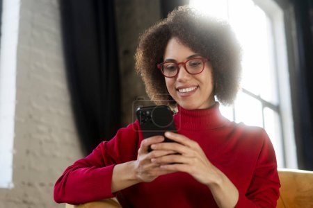 Téléchargez les photos : Belle femme afro-américaine souriante tenant un téléphone portable regardant des vidéos assis à la maison. Femme moderne portant des lunettes rouges à l'aide d'une application mobile, faire des achats en ligne, commander de la nourriture. Services bancaires mobiles - en image libre de droit