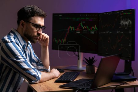 Night portrait of pensive trader looking at laptop monitor, working online with crypto chart in modern office. Online trading, successful business concept