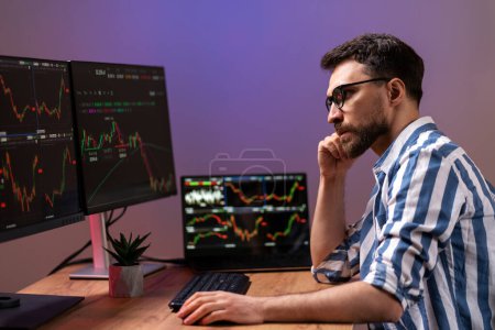Serious handsome man, crypto trader looking at computer monitor, working online with crypto chart sitting in modern office. Broker research stock market. Successful business, online trading concept