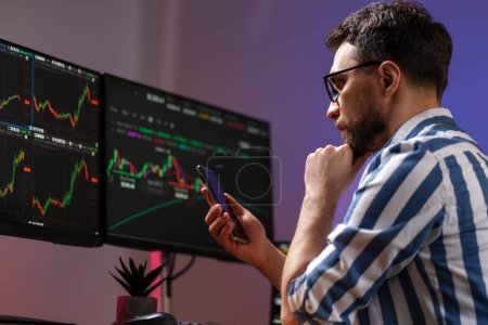 Night portrait of serious handsome trader holding mobile phone, using laptop computer working online with crypto chart sitting in modern office. Successful business, online trading concept