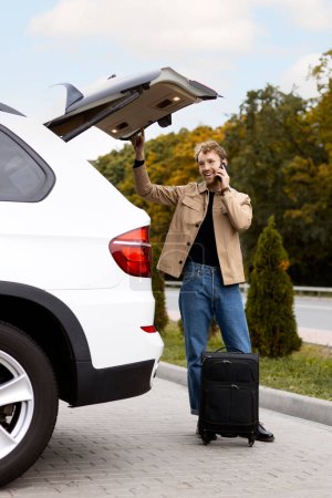 Photo for Smiling man standing near car, talking on mobile phone. Young man pulling out suitcase from trunk of car - Royalty Free Image