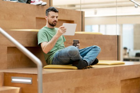 Photo for Pensive student studying holding digital tablet in university campus, online education concept. Modern latin guy reading e book, watching videos, holding coffee cup sitting in lotus pose - Royalty Free Image