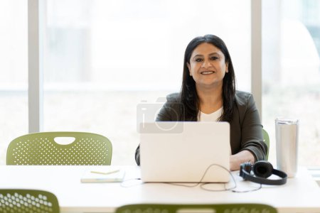 Photo for Smiling Indian manager using laptop computer looking at camera working in office. Portrait of confident successful secretary sitting at workplace - Royalty Free Image