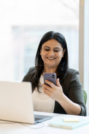Photo for Smiling Indian business woman using laptop, holding mobile phone, checking email sitting in modern office. Portrait of successful asian secretary sitting at workplace. Technology concept - Royalty Free Image