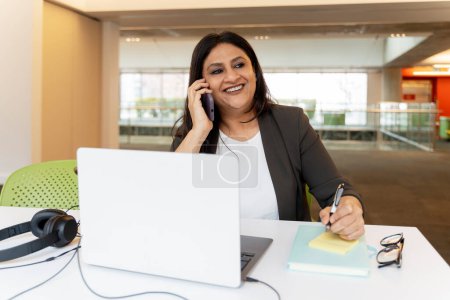 Photo for Confident smiling Indian businesswoman talking on mobile phone, using laptop, working online in modern office. Happy mature secretary answering call, taking notes at workplace. Successful business - Royalty Free Image