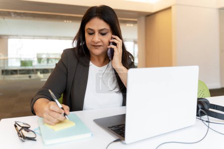 Téléchargez les photos : Femme indienne mature pensive parlant téléphone mobile, prenant des notes, utilisant un ordinateur portable, travaillant au bureau. Gestionnaire asiatique occupé projet de planification, remue-méninges au lieu de travail. Concept multitâche - en image libre de droit