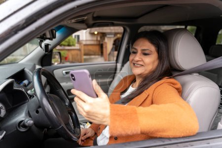 Foto de Confiado sonriendo mujer india madura sosteniendo el teléfono móvil, comprobando el correo electrónico sentado dentro de coche nuevo. Transporte, concepto de conducción distraído - Imagen libre de derechos