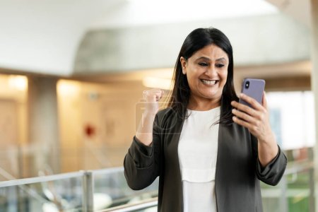 Foto de Mujer madura india feliz comprueba el teléfono móvil, cierra el puño, se siente feliz ganar licitación, se regocija en el acuerdo exitoso, sonríe, de pie en el interior de la oficina moderna. Gente. Negocios. Concepto tecnológico - Imagen libre de derechos