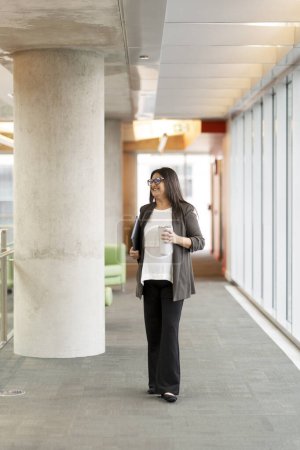 Photo for Full length portrait of confident Asian adult woman, entrepreneur, office worker, sales manager, business owner, carrying a laptop while walking in the corporate office with a thermo bottle in hands - Royalty Free Image