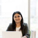Confident portrait of a mature Indian woman in formal suit, sitting at desk with laptop, smiling a cheerful toothy smile and confidently looking at camera. Businesswoman at workplace in modern office