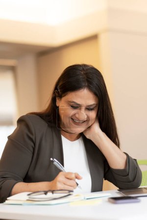 Foto de Sonriente exitosa madura mujer de negocios asiática, empresario, oficinista, gerente haciendo notas, escritura a mano en un cuaderno mientras está sentado en el escritorio con el ordenador portátil. Concepto de personas y negocios - Imagen libre de derechos