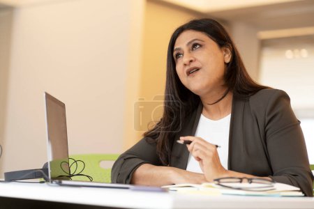 Photo for Multitasking Asian mature businesswoman sitting at desk, working online on laptop, thoughtfully looking aside while planning new business project in office interior. People Technology Business concept - Royalty Free Image
