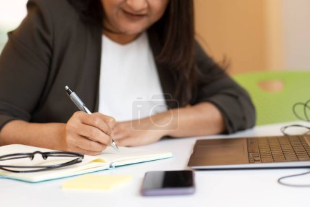 Téléchargez les photos : Focus sélectif sur la main de la femme d'affaires asiatique, prendre des notes, écrire à la main sur un ordinateur portable tout en étant assis au bureau avec ordinateur portable et appareils électroniques numériques. Concept de personnes et d'entreprises - en image libre de droit
