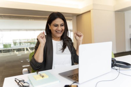 Photo for Mature charming successful Indian woman sitting at a desk with laptop in modern office interior, smiling, clenching fists with happiness, rejoicing in the conclusion of a deal or winning a tender. - Royalty Free Image