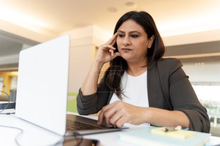 Photo for Puzzled thoughtful Asian mature woman entrepreneur, reflecting on own business project, sitting at desk with laptop while working online in a modern office. Portrait of successful motivated people - Royalty Free Image