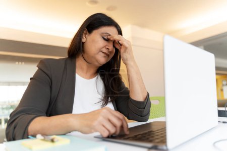 Foto de Mujer madura asiática estresada sosteniendo la cabeza en las manos, sintiéndose desmotivada mientras está sentada en el escritorio de la oficina y trabajando en línea en el portátil. Retrato del exhausto director ejecutivo multitarea en el lugar de trabajo - Imagen libre de derechos