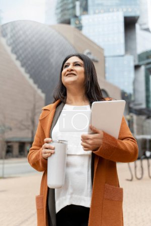 Photo for Stylish elegant confident positive mature Asian woman in orange casual suit, holding digital tablet while walking and standing in city centre on urban street. People, business and technology concept - Royalty Free Image