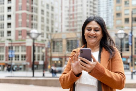 Téléchargez les photos : Sourire positif femme indienne mature, en utilisant un smartphone, ajuste le navigateur sur les applications mobiles tout en marchant dans la rue urbaine, vérifie le contenu des médias sociaux, naviguant sur les sites Web. Personnes Technologie de style de vie - en image libre de droit
