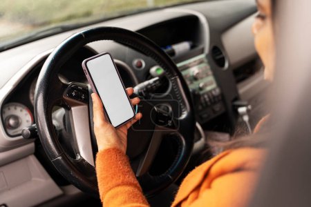 Detalles en smartphone con pantalla blanca en blanco con espacio libre para insertar su aplicación móvil, en la mano de una conductora, sentada en el asiento del conductor en un coche moderno. Gente. Tecnología. Transportes