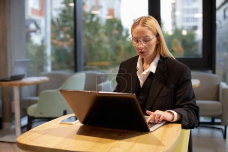 Téléchargez les photos : Femme d'affaires pensive portant des lunettes à l'aide d'un ordinateur portable travaillant en ligne dans un bureau moderne. Rédacteur pigiste tapant sur le clavier, projet de planification sur le lieu de travail. Une entreprise prospère - en image libre de droit