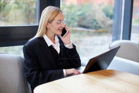 Photo for Young business woman wearing formal suit talking on mobile, using laptop in modern office - Royalty Free Image