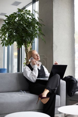 Photo for Beautiful confident blonde saleswoman talking on mobile phone, using laptop computer working in modern office. Attractive middle aged secretary wearing stylish suit answering call sitting at workplace - Royalty Free Image