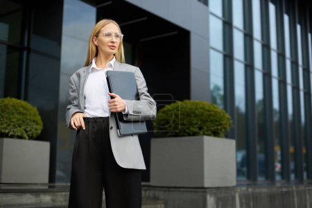 Photo for Confident blonde businesswoman wearing stylish eyeglasses, holding laptop computer looking away standing on the street, copy space. Successful business - Royalty Free Image