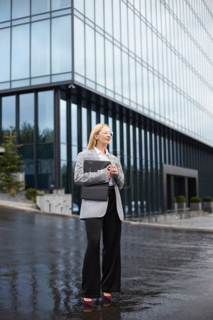 Foto de Pensativa mujer de negocios rubia de mediana edad sosteniendo portátil, mirando hacia otro lado, esperando a alguien en la calle, día lluvioso - Imagen libre de derechos