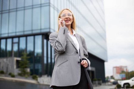 Foto de Retrato de la sonriente mujer de negocios rubia confiada que lleva gafas con estilo hablando en el teléfono móvil de pie en la calle urbana. Tecnología, concepto de negocio exitoso - Imagen libre de derechos