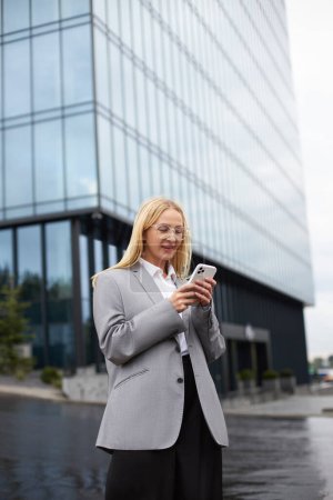 Photo for Confident smiling businesswoman wearing stylish eyeglasses using mobile phone reading text message, check email, communication online standing on the street. Technology concept - Royalty Free Image