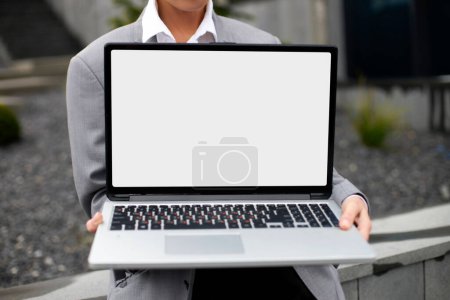 Photo for Cropped view of the business woman wearing formal suit holding laptop computer with blank white screen at the street, mockup - Royalty Free Image