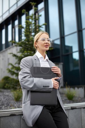Photo for Middle aged businesswoman wearing stylish eyeglasses, holding laptop looking away on the street - Royalty Free Image