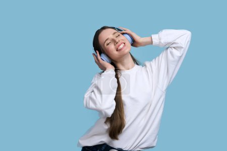 Attraktive kaukasische brünette Frau mit drahtlosen Kopfhörern, die einen coolen Track hört und sich zu Musik bewegt, isoliert vor blauem Hintergrund. Menschen, Tanz, Hobby, Unterhaltung und Lifestylekonzept