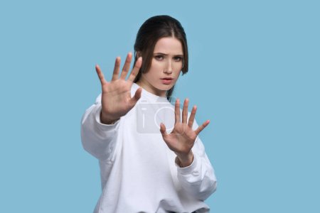 Photo for Worried anxious Caucasian young woman in white sweatshirt, showing stop sign with palms facing camera, expressing misunderstanding, disappointment, denial and rejection over isolated blue background - Royalty Free Image