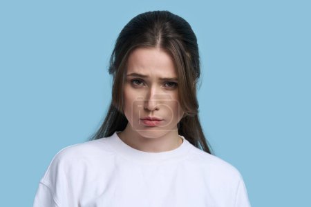 Photo for Charming Caucasian young brunette woman in white sweatshirt, looking pensively at camera, pouting lips, expressing misunderstanding and sadness, isolated on blue background. People Lifestyle Emotions - Royalty Free Image