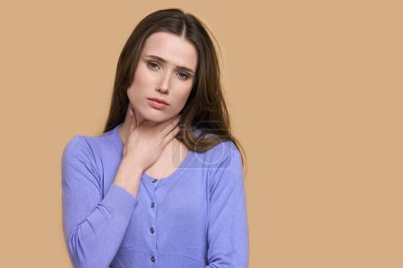 Photo for Young woman looking sick, touching painful neck, sore throat for flu, cold and viral infection, isolated on beige background. Young woman touching the neck feeling unwell, coughing, looking at camera - Royalty Free Image