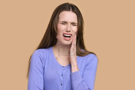 Photo for Frustrated Caucasian young woman, holding hand on her cheek, screaming, crying, suffering from awful toothache, isolated on beige background. Dentistry. Dental health concept. Healthcare and medicine - Royalty Free Image
