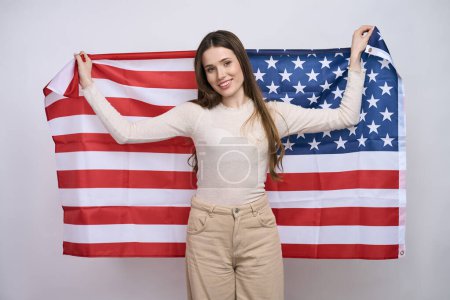 Foto de Mujer bonita feliz en ropa casual, llevando bandera americana, sonriendo mirando a la cámara, aislada sobre fondo blanco. 4 de julio. Concepto del Día del Patriotismo y la Independencia - Imagen libre de derechos