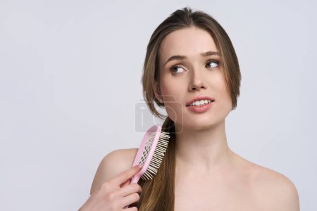 Photo for Attractive young Caucasian woman, combing beautiful long shiny silky hair with a pink hair brush, smiling, looking aside, posing with naked shoulders, isolated on white background. Copy space for ads - Royalty Free Image