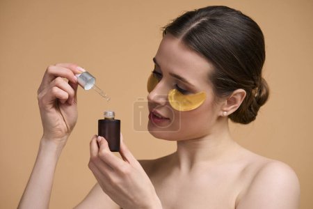 Téléchargez les photos : Portrait rapproché de belle jeune femme caucasienne, avec des patchs d'oeil d'hydrogel doré, tenant une bouteille avec du sérum d'acide hyaluronique anti-vieillissement, posant les épaules nues, isolé sur fond beige - en image libre de droit