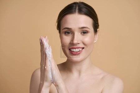 Photo for Close-up beauty portrait of Caucasian confident beautiful young woman 20-30 years old, with cleansing soap foam or makeup remover on hands, smiling looking at camera, isolated over beige background - Royalty Free Image