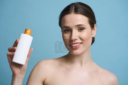 Photo for Caucasian attractive young woman with healthy glowing skin, holding white bottle with sunscreen lotion, smiling looking at camera, posing bare shoulders, isolated on blue background. Skin care concept - Royalty Free Image